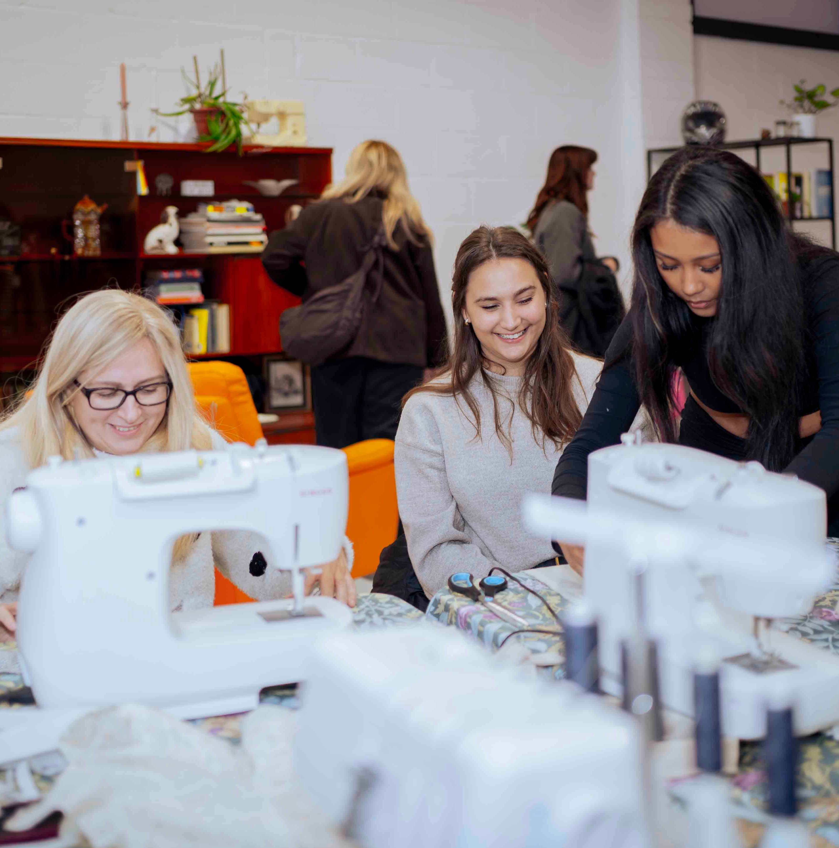 Three women gathered around sewing machines in a bright indoor workshop or classroom setting, with shelves and other people visible in the background.