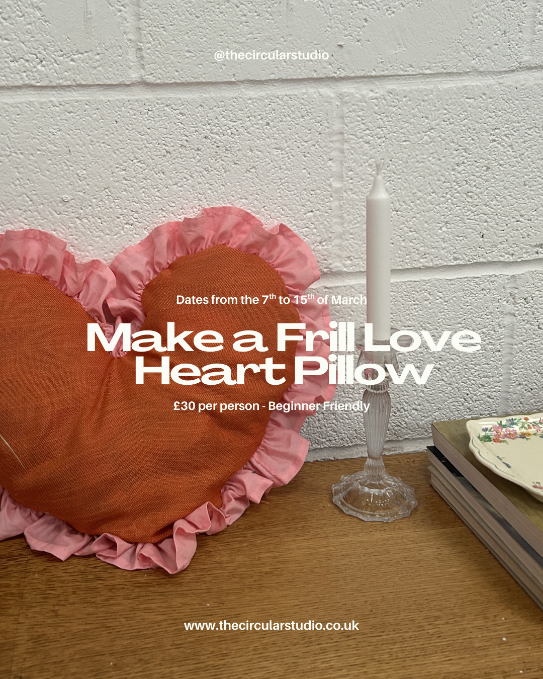 Heart-shaped pillow with frills on a wooden surface next to a candle, against a white brick wall.