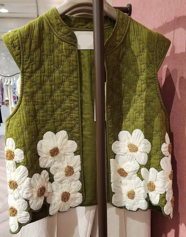 An olive green quilted vest with large white daisy embroidery on the hem, hanging on a clothing rack in a boutique.