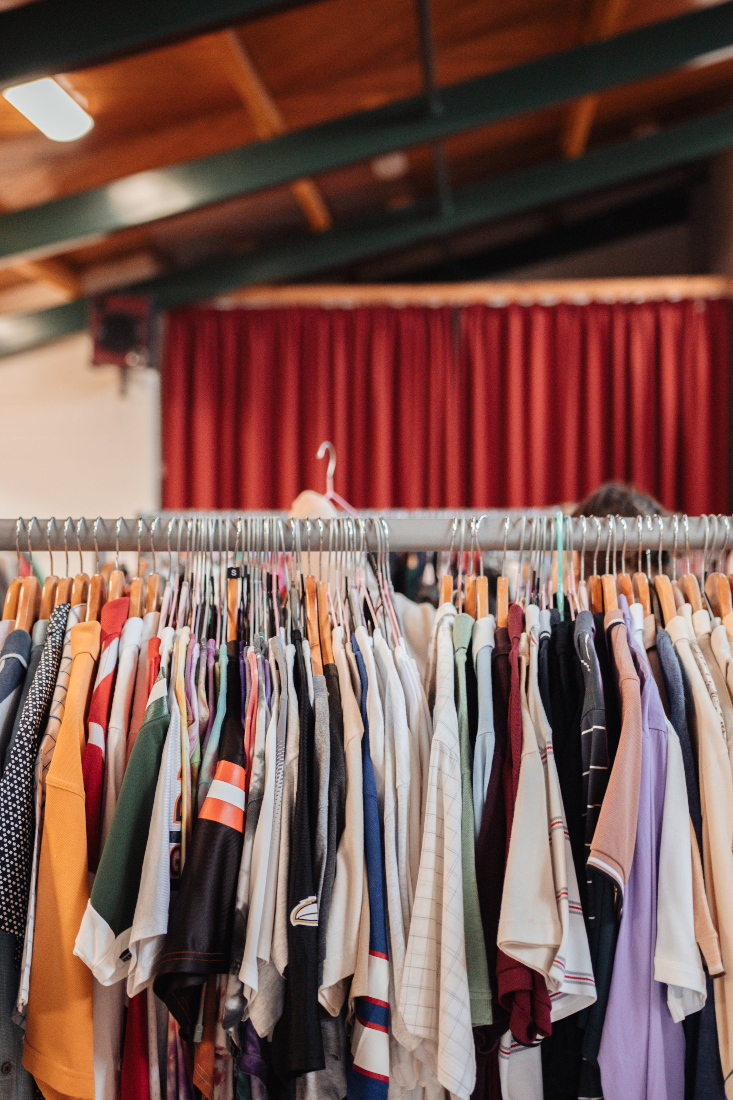 A clothing rack filled with assorted vintage garments in various colors, set in a room with a red curtain and wooden ceiling beams in the background.