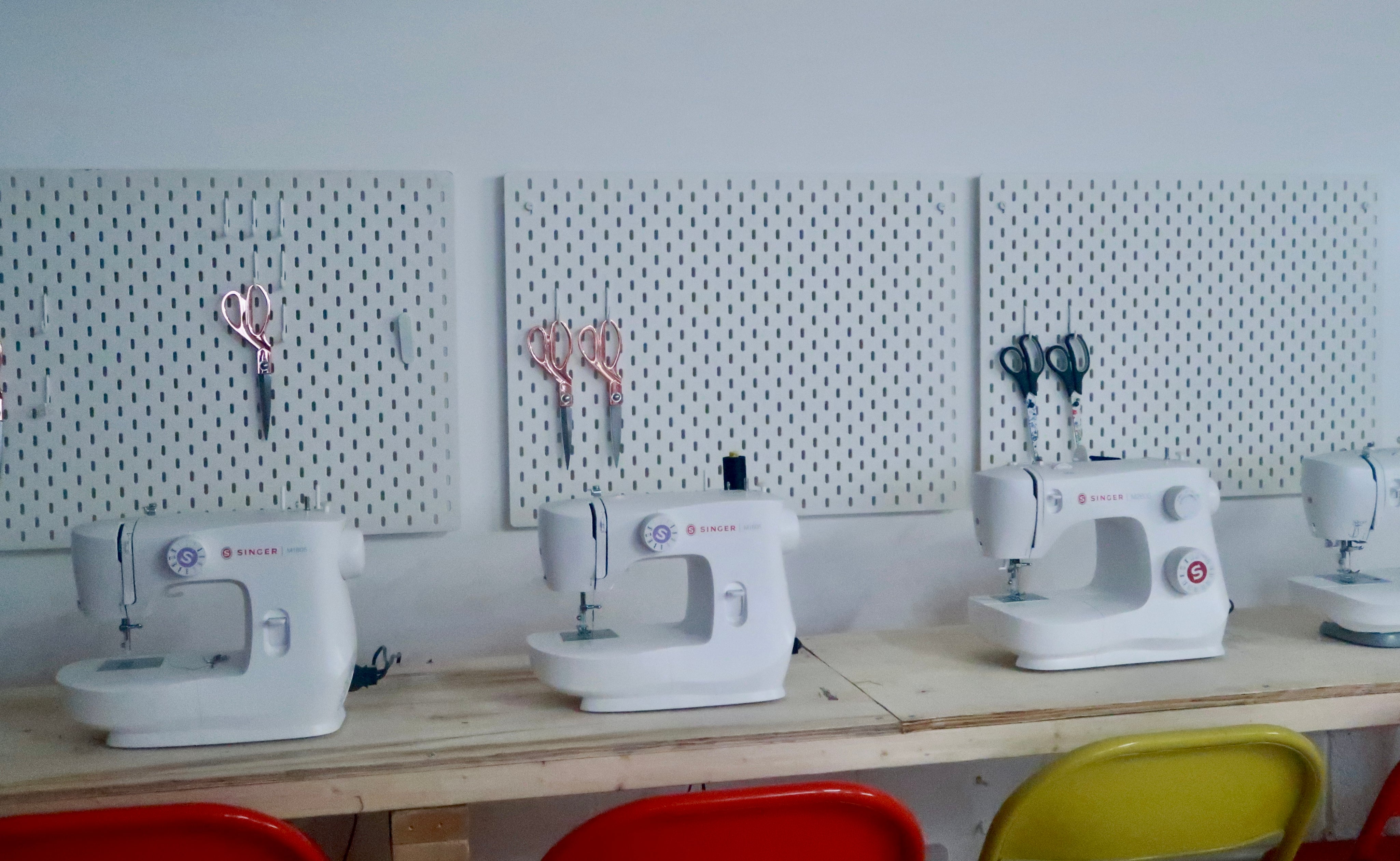 A row of white sewing machines on a wooden table in a bright room, with pegboards on the wall holding scissors and colorful chairs in front.