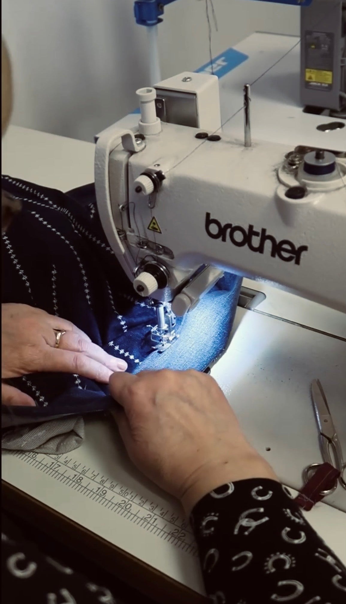 A close-up of a Brother industrial sewing machine in use, with a person's hands guiding blue patterned fabric under the needle. The sewing machine is white with black branding, and a pair of scissors rests on the table.
