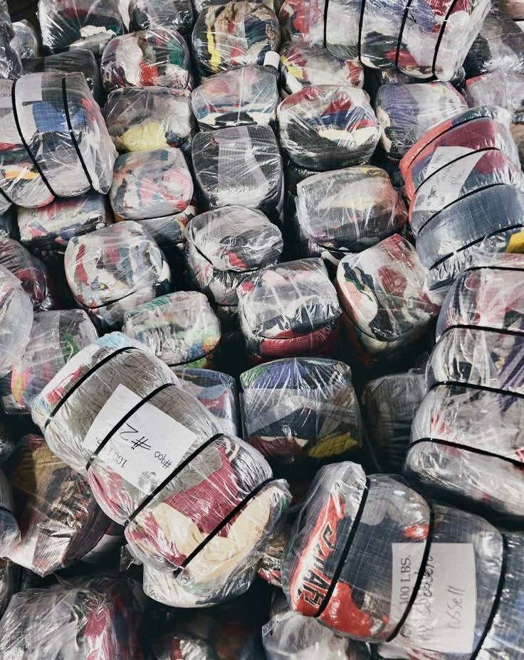 A dense pile of bundled clothing parcels wrapped in clear plastic with black straps and white labels, stacked tightly together.