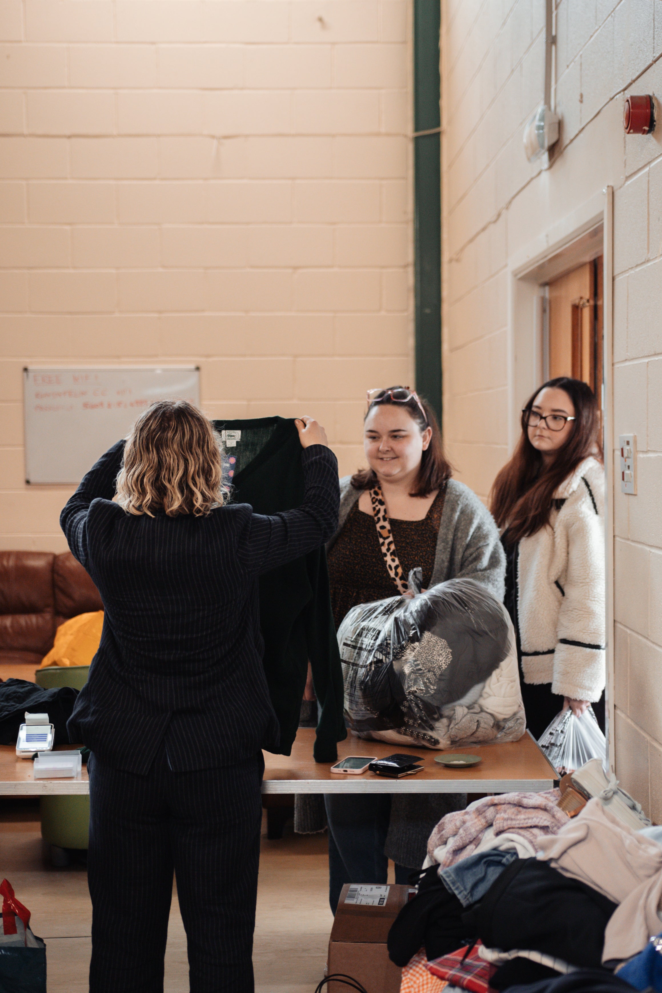 A woman in a black outfit holds up a black garment in a room with beige brick walls, while two other women stand nearby at a table with folded clothes.